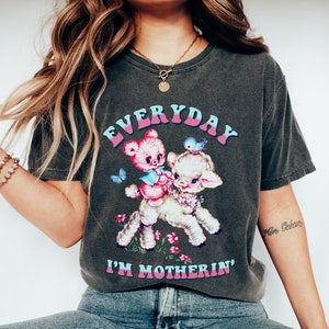 Op de afbeelding: Een donkergrijs T-shirt met een vintage-stijl afbeelding van een teddybeer die op een lammetje rijdt. De tekst "EVERYDAY I'M MOTHERIN'" is in roze en blauwe letters op het shirt gedrukt.