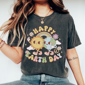 May include: A dark grey t-shirt with a graphic of a smiling sun and earth holding hands. The graphic is surrounded by a rainbow, clouds, flowers, and the text "Happy Earth Day".