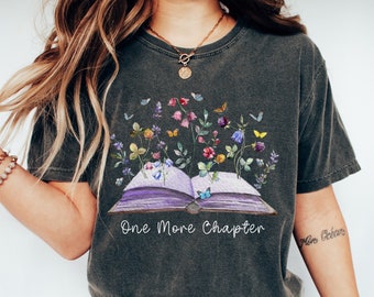 May include: A dark gray t-shirt with a graphic of an open book with flowers and butterflies growing from it. The text "One More Chapter" is printed below the book.