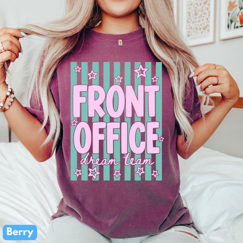 K&ouml;nnte beinhalten: Ein beerenfarbenes Kurzarm-T-Shirt mit einem Grafikdruck aus vertikalen blaugr&uuml;nen Streifen. Der Text lautet FRONT OFFICE in gro&szlig;en rosa Buchstaben, darunter steht dream team in Schreibschrift. Kleine rosa Sterne umgeben den Text.
