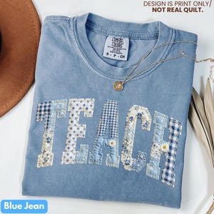 May include: A blue jean Comfort Colors t-shirt with the word "TEACH" appliqued in various blue patterned fabrics. A gold necklace with a pendant rests on the shirt. The text "DESIGN IS PRINT ONLY/ NOT REAL QUILT" is printed at the top.