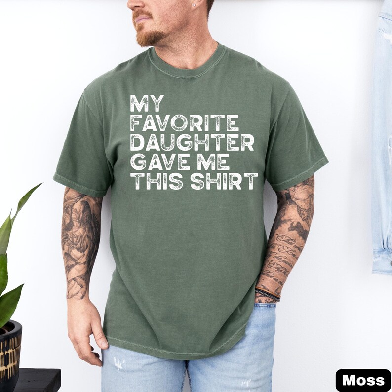 Pu&ograve; includere: Una maglietta verde muschio con la scritta "MY FAVORITE DAUGHTER GAVE ME THIS SHIRT" in lettere bianche. La maglietta &egrave; di colore pieno e ha maniche corte. La maglietta &egrave; indossata da una persona.