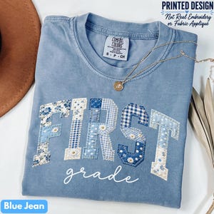 May include: A blue jean colored t-shirt with a printed design featuring the word FIRST in large block letters filled with various blue and white floral and gingham patterns, and the word grade in white script below the block letters.