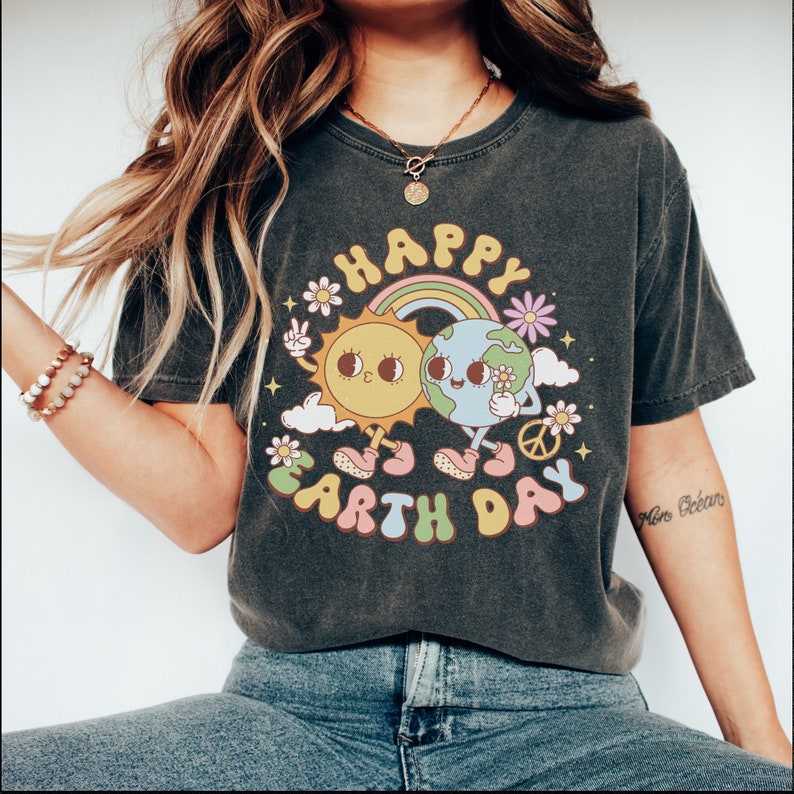 May include: A dark grey t-shirt with a graphic of a smiling sun and earth holding hands. The graphic is surrounded by a rainbow, clouds, flowers, and the text "Happy Earth Day".