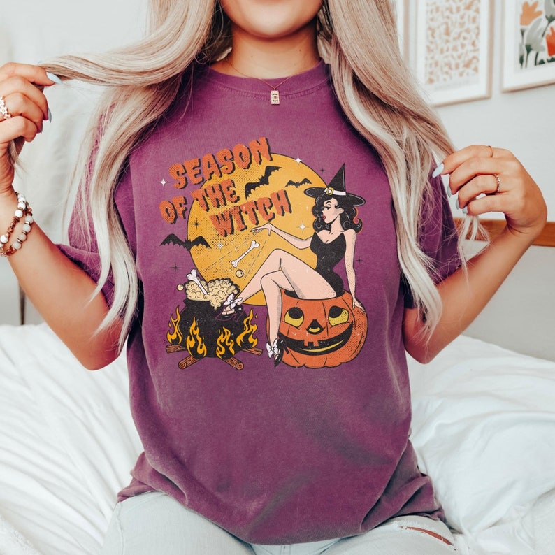 May include: Purple t-shirt featuring a vintage illustration of a witch sitting in a pumpkin. The text "Season of the Witch" is printed on the shirt.