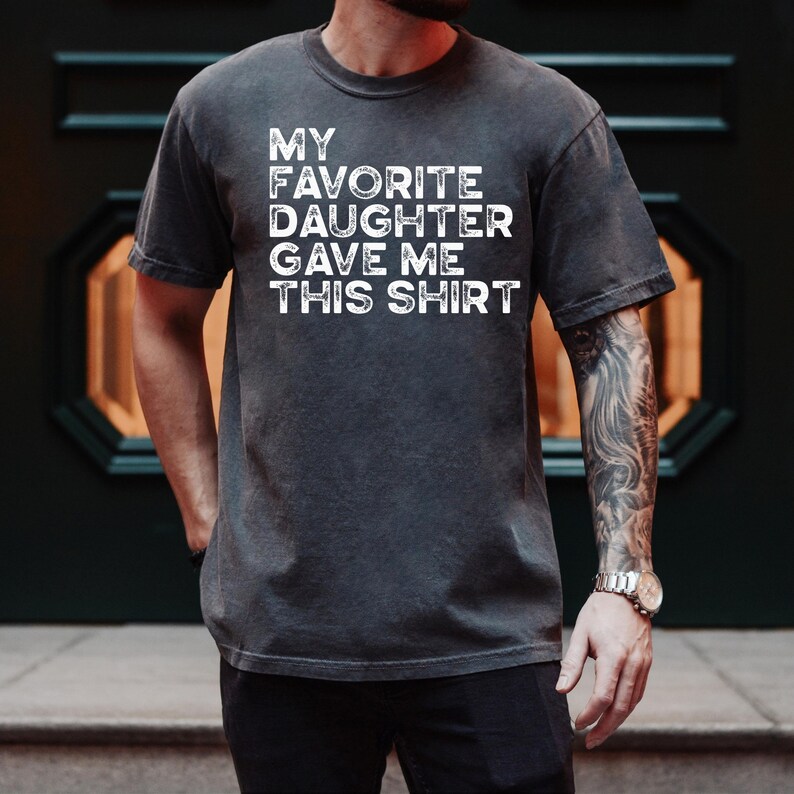 Pu&ograve; includere: T-shirt grigio scuro con la scritta bianca "MY FAVORITE DAUGHTER GAVE ME THIS SHIRT". La maglietta ha maniche corte e girocollo. Il tessuto appare leggermente sbiadito.
