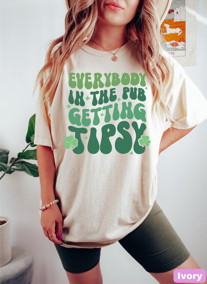 Puede incluir: Camiseta color marfil con la frase "EVERYBODY IN THE PUB GETTING TIPSY" en tonos verdes, adornada con tr&eacute;boles y estrellas. La camiseta tiene un corte holgado y oversize.