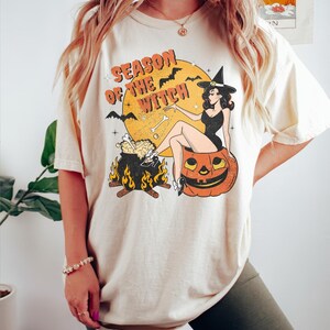 May include: A cream coloured t-shirt with a vintage graphic of a witch sitting on a pumpkin. The graphic includes the text "Season of the Witch" and features a cauldron with flames and bats.