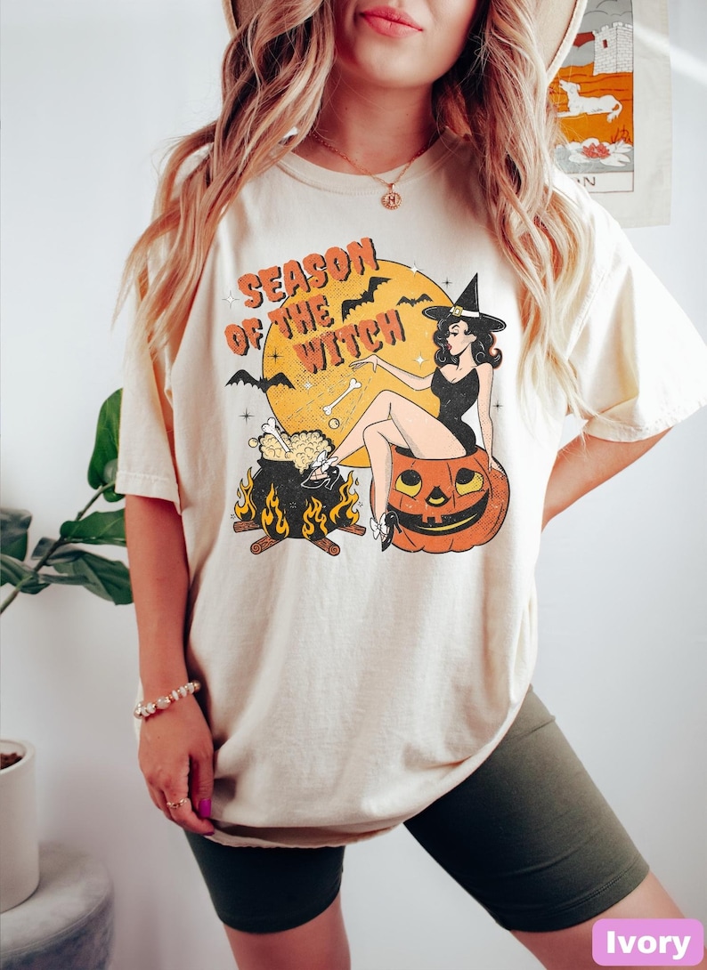 May include: A cream coloured t-shirt with a vintage graphic of a witch sitting on a pumpkin. The graphic includes the text "Season of the Witch" and features a cauldron with flames and bats.