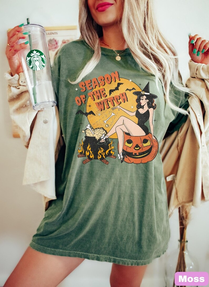 May include: Green t-shirt featuring a vintage-style graphic of a witch sitting on a pumpkin. The graphic reads "Season of the Witch".