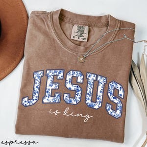 May include: A brown t-shirt with a white floral design that says "JESUS is king" in blue letters.