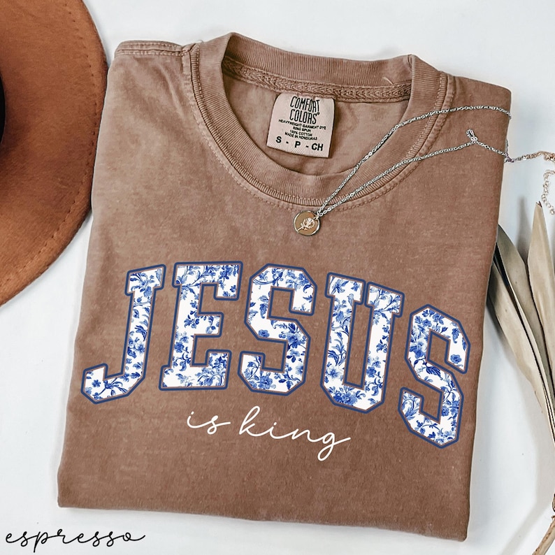 May include: A brown t-shirt with a white floral design that says "JESUS is king" in blue letters.