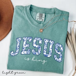 May include: A light green t-shirt with the words "Jesus is king" in white and blue floral lettering. The shirt is made of a soft, comfortable material.