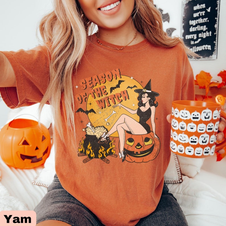 May include: A burnt orange t-shirt featuring a vintage illustration of a witch sitting in a pumpkin. The witch is wearing a black dress and a witch's hat. The text "Season of the Witch" is printed on the shirt.