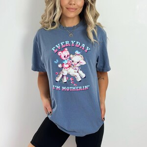 Op de afbeelding: Een blauw t-shirt met een afbeelding van een teddybeer die op een lammetje rijdt. De tekst op het shirt luidt "EVERYDAY I'M MOTHERIN'".