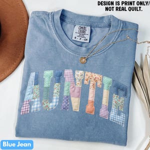 May include: A blue jean colored t-shirt with the word "AUNTIE" in patchwork letters. The letters are made of various floral and patterned fabrics. A gold necklace with a pendant is draped across the shirt. The text "DESIGN IS PRINT ONLY/ NOT REAL QUILT" is at the top.