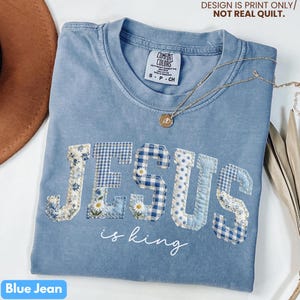May include: A blue jean colored t-shirt with the word "JESUS" in patchwork letters, featuring floral, gingham, and polka dot patterns. The phrase "is king" is written below in script. A gold necklace with a pendant is draped across the shirt.