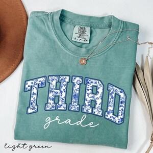 K&ouml;nnte beinhalten: Ein hellgr&uuml;nes T-Shirt mit einem blauen und wei&szlig;en Blumenmuster, auf dem in marineblau und wei&szlig; "THIRD grade" steht.
