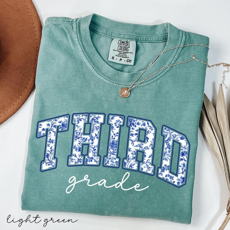 K&ouml;nnte beinhalten: Ein hellgr&uuml;nes T-Shirt mit einem blauen und wei&szlig;en Blumenmuster, auf dem in marineblau und wei&szlig; "THIRD grade" steht.