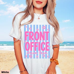 K&ouml;nnte beinhalten: Ein wei&szlig;es kurz&auml;rmeliges T-Shirt mit einem Grafikdruck aus hellblauen vertikalen Streifen. Der Text FRONT OFFICE ist in gro&szlig;en rosa Buchstaben gedruckt, darunter stehen die Worte dream team in rosa Schreibschrift, umgeben von kleinen rosa Sternen.