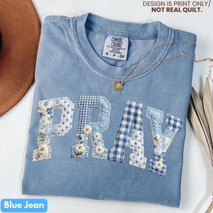 May include: A blue jean colored t-shirt with the word "PRAY" appliqued across the front in a patchwork design. The letters feature floral, polka dot, and gingham patterns. A gold necklace with a pendant rests on the shirt. The text "DESIGN IS PRINT ONLY/ NOT REAL QUILT" is at the top.