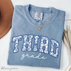 K&ouml;nnte beinhalten: Ein hellblaues T-Shirt mit einem wei&szlig;en Blumenmuster, auf dem in einer blauen und wei&szlig;en Musterfont "THIRD grade" steht.