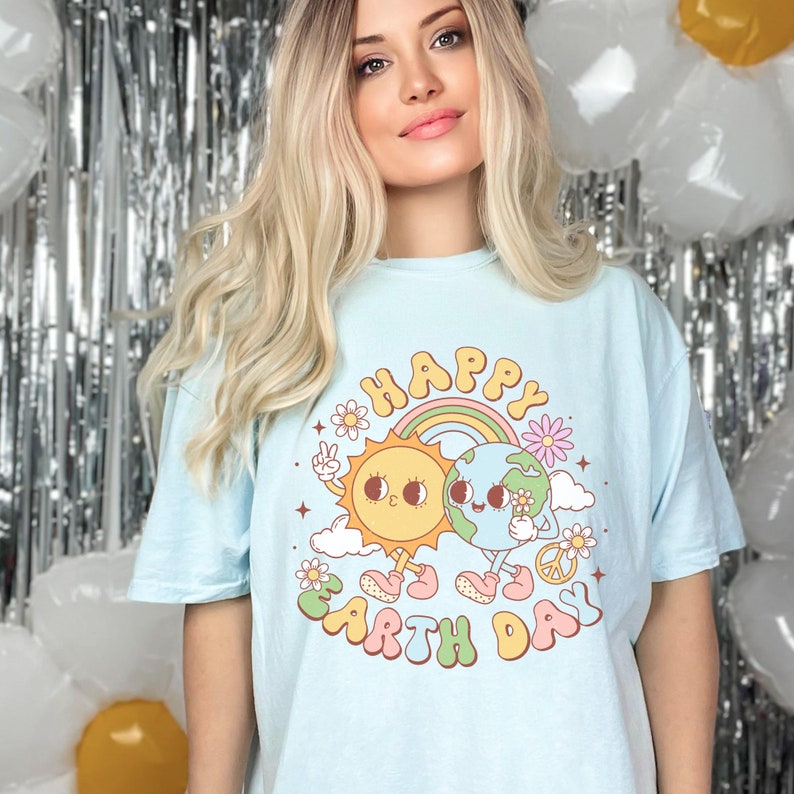 May include: A light blue t-shirt with a graphic of a smiling sun and earth holding hands. The graphic includes a rainbow, clouds, and flowers. The text "Happy Earth Day" is written in a colorful, retro font.