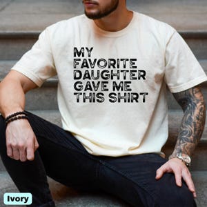 Pu&ograve; includere: T-shirt color avorio con la scritta "MY FAVORITE DAUGHTER GAVE ME THIS SHIRT" in lettere nere. La maglietta &egrave; a maniche corte e sembra essere fatta di un materiale morbido.
