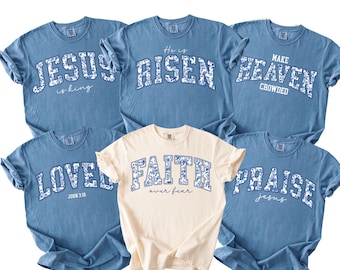 May include: A selection of blue and cream-coloured t-shirts. Each shirt displays a religious phrase in a floral design. Phrases include "Jesus is King," "He is Risen," and "Make Heaven Crowded."