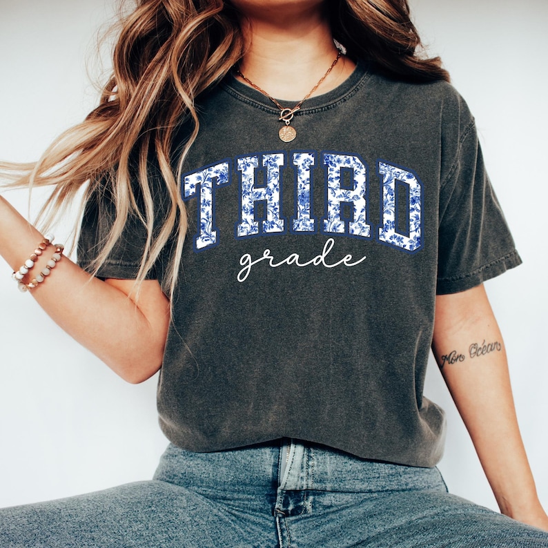K&ouml;nnte beinhalten: Ein dunkelgraues T-Shirt mit einem blauen und wei&szlig;en Blumenmuster, auf dem in wei&szlig;er Schrift "THIRD grade" steht.