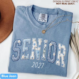 May include: A blue jean colored t-shirt with the word "SENIOR" in a patchwork design, with the year "2027" below. The letters have floral and gingham patterns. A gold necklace with a pendant rests on the shirt. The text "DESIGN IS PRINT ONLY/ NOT REAL QUILT" is at the top.