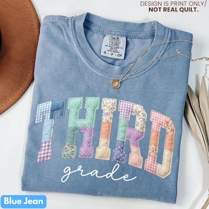 May include: Blue jean colored t-shirt with the words "THIRD grade" in a patchwork design. The shirt has a crew neck and short sleeves. A gold necklace with a pendant is draped across the shirt. The text "DESIGN IS PRINT ONLY/ NOT REAL QUILT" is at the top.
