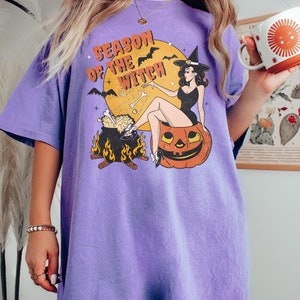 May include: Purple t-shirt featuring a graphic of a witch sitting on a pumpkin with the text "Season of the Witch".