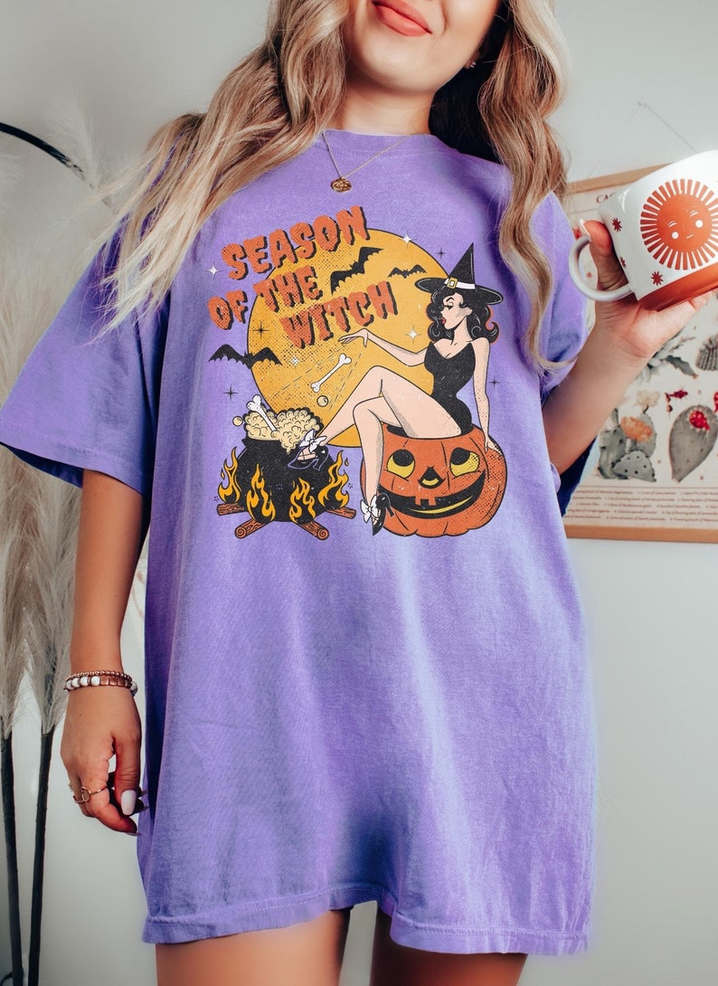 May include: Purple t-shirt featuring a graphic of a witch sitting on a pumpkin with the text "Season of the Witch".