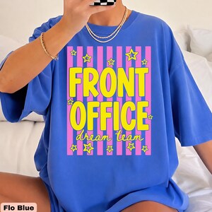 K&ouml;nnte beinhalten: Ein hellblaues Kurzarm-T-Shirt mit einem Grafikdruck aus vertikalen rosa Streifen und gelben Sternen. Der Text FRONT OFFICE ist in gro&szlig;en gelben Buchstaben gedruckt, darunter steht dream team in kleinerer gelber Schreibschrift.