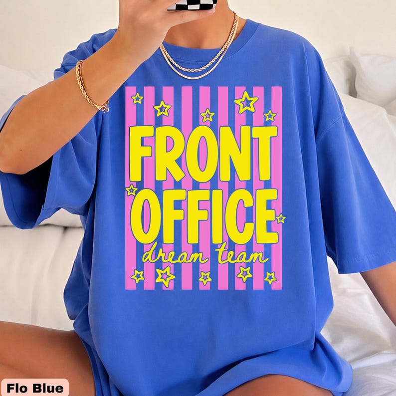 K&ouml;nnte beinhalten: Ein hellblaues Kurzarm-T-Shirt mit einem Grafikdruck aus vertikalen rosa Streifen und gelben Sternen. Der Text FRONT OFFICE ist in gro&szlig;en gelben Buchstaben gedruckt, darunter steht dream team in kleinerer gelber Schreibschrift.