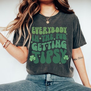 Puede incluir: Camiseta gris oscuro con la frase "EVERYBODY IN THE PUB GETTING TIPSY" en tonos verdes. El dise&ntilde;o incluye tr&eacute;boles y estrellas, ideal para las celebraciones del D&iacute;a de San Patricio.