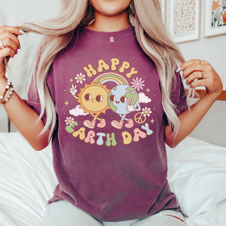 May include: Purple t-shirt with a graphic of a smiling sun and earth holding a flower. The graphic says "Happy Earth Day" in rainbow letters.