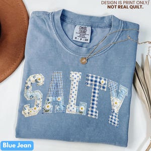 May include: A blue jean colored t-shirt with the word "SALTY" appliqued across the front. The letters are made of various blue patterned fabrics, including floral and gingham designs. A gold necklace with a pendant is draped across the shirt.