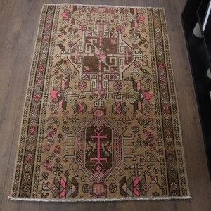 Op de afbeelding: Een rechthoekig, beige tapijt met een gedetailleerd, geometrisch patroon in bruin, roze en groen. Het tapijt heeft een centraal medaillonontwerp en een rand met herhaalde bloemmotieven. Het ligt op een houten vloer.