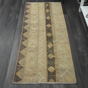 Puede incluir: Una alfombra de pasillo beige y marrón con un patrón geométrico. La alfombra tiene un aspecto descolorido y está hecha de un material suave y tejido.