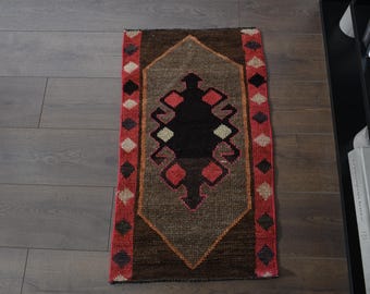 Puede incluir: Una alfombra rectangular estampada con un diseño geométrico central en negro, rojo y beige. La alfombra tiene un borde marrón y está colocada sobre un suelo de madera. El diseño de la alfombra presenta formas de diamante y flecha.