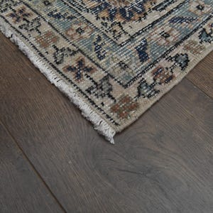 May include: Close-up of the fringe edge of a beige rug with a blue and brown floral pattern. The rug is on a dark brown wood floor.