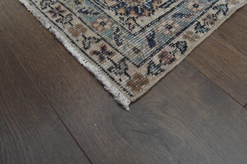 May include: Close-up of the fringe edge of a beige rug with a blue and brown floral pattern. The rug is on a dark brown wood floor.