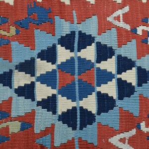 K&ouml;nnte beinhalten: Eine Nahaufnahme eines bunten gewebten Teppichs mit einem geometrischen Muster in Rot-, Blau-, Wei&szlig;- und Beiget&ouml;nen. Der Teppich hat eine zentrale Diamantform mit komplizierten Details.