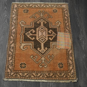 May include: A small, rectangular rug with a brown and orange geometric pattern. The rug has a fringe border and a small patch of fabric sewn on the right side.