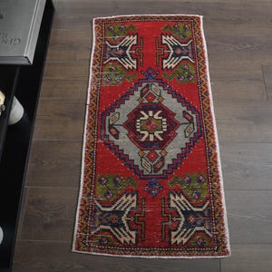 Puede incluir: Una alfombra rectangular, tejida a mano, con un fondo rojo y patrones geométricos intrincados en gris, azul, verde y crema. La alfombra presenta un diseño central de diamante y bordes con motivos repetidos. La alfombra está colocada sobre un suelo de madera oscura.