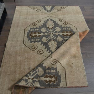 Op de afbeelding: Beige en grijs patroonkleed met een geometrisch ontwerp. Het kleed heeft een centraal medaillon en randpatronen in grijstinten en bruin. Het kleed is gedeeltelijk opgevouwen en onthult de onderkant.
