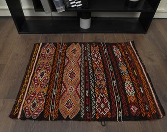 May include: A traditional black, red, orange, and white patterned rug with geometric designs. The rug is made of wool and has a fringed edge.