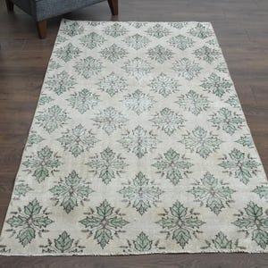 Puede incluir: Alfombra rectangular con un patrón floral repetido en tonos verdes y grises sobre un fondo crema. La alfombra está colocada sobre un suelo de madera oscura, mostrando su diseño y textura. Un elemento de decoración del hogar.